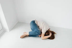 Persona tumbada en el suelo, encogida, brazos cubriendo la cabeza. Respira agitado, cuerpo tenso. Alrededor, una habitación vacía. No hay nadie ni nada. Solo el piso frío y el miedo apretando desde adentro.