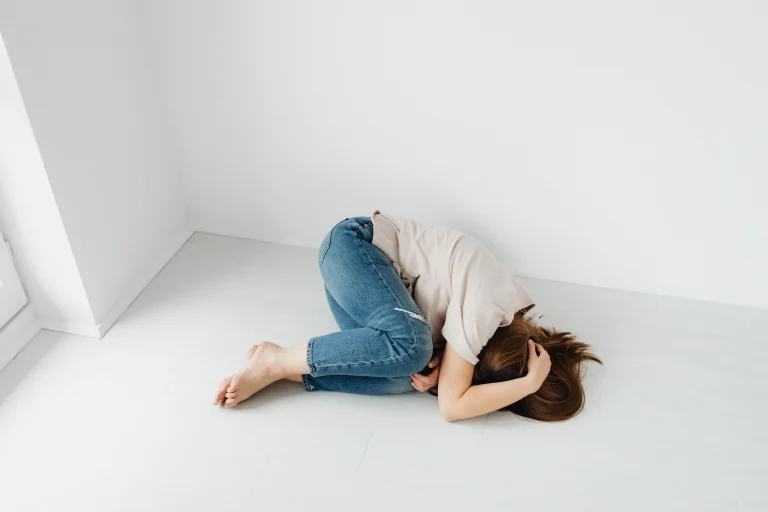 Persona tumbada en el suelo, encogida, brazos cubriendo la cabeza. Respira agitado, cuerpo tenso. Alrededor, una habitación vacía. No hay nadie ni nada. Solo el piso frío y el miedo apretando desde adentro.